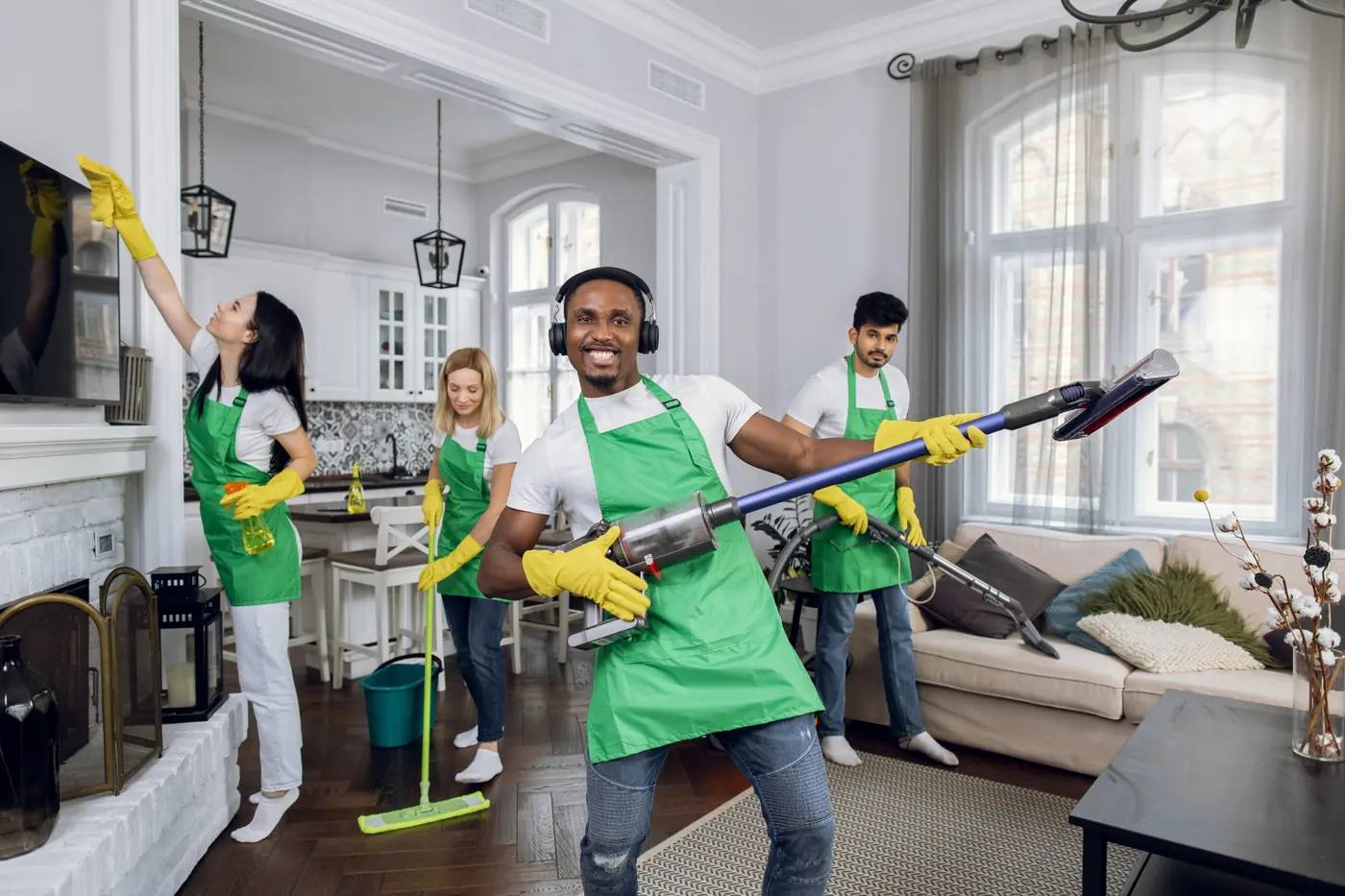 A cleaning crew in green aprons and yellow gloves dusting, mopping and vacuuming a bright living room