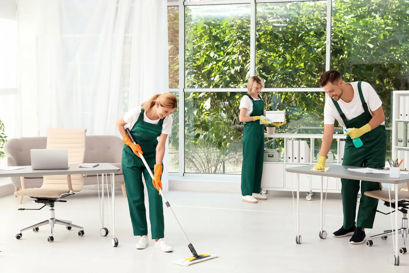 A cleaning crew in green coveralls mopping the floor and wiping desks in a bright, modern office with large windows