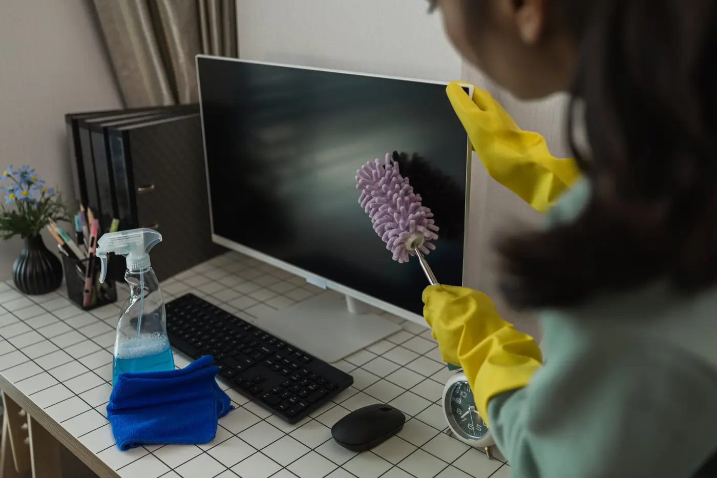 A cleaner in yellow gloves dusting a computer monitor on a tidy office desk