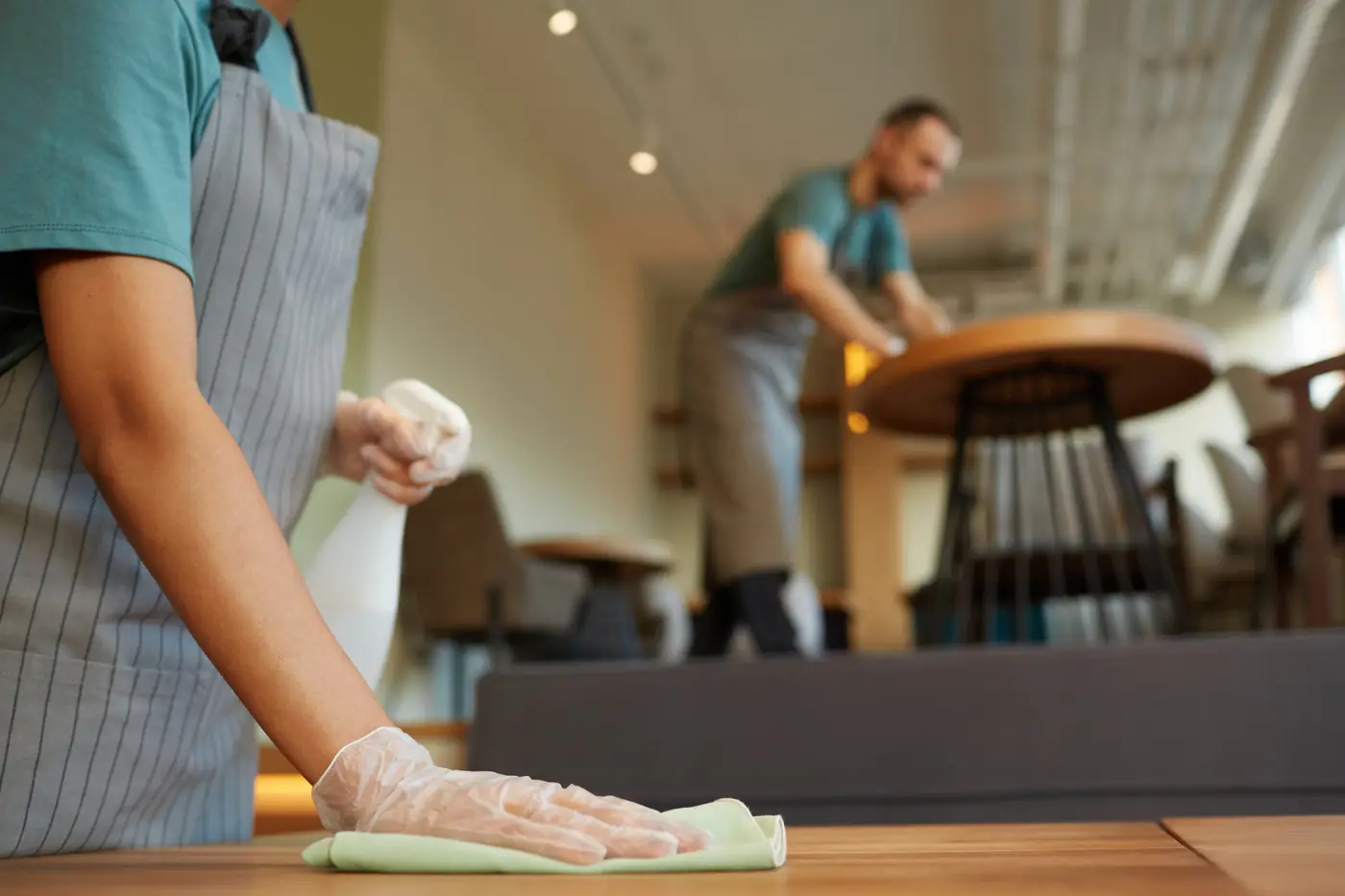 A cleaner in disposable gloves wiping a wooden restaurant table with a spray bottle and cloth