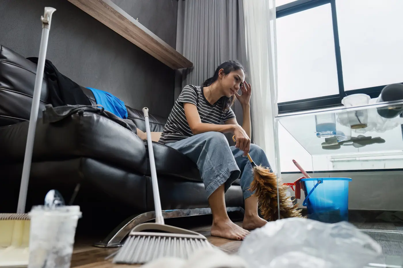 A person sitting on a sofa surrounded by a broom, bucket and duster in a cluttered living room