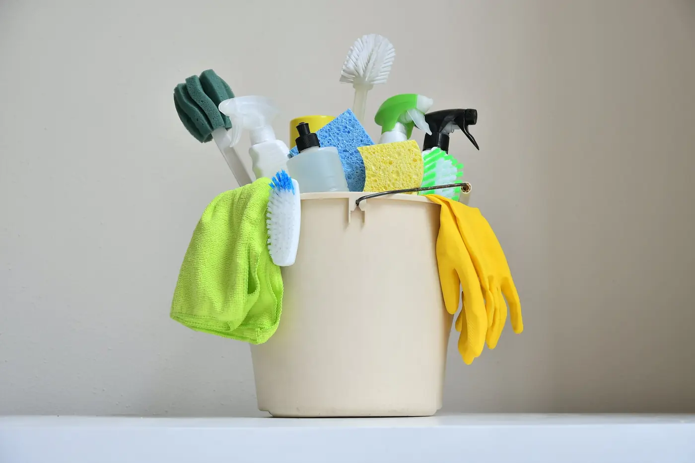 A bucket of cleaning supplies with brushes, sponges, spray bottles, a microfiber cloth and rubber gloves
