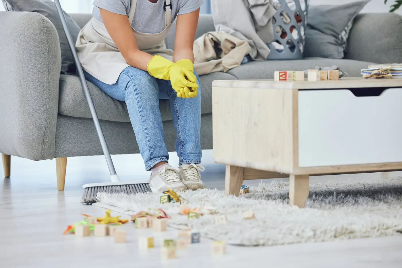 A person in yellow gloves resting on a sofa beside a broom, with toys scattered across the living room floor